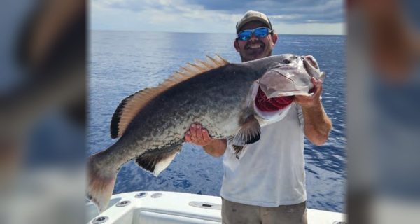 Capt. Chris Endicott in a cap and blue mirrored sunglasses holding up a huge grouper on the deck of his boat, with the open ocean behind him