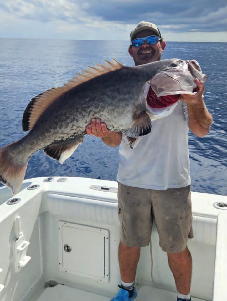 Capt. Chris Endicott in a cap and blue mirrored sunglasses holding up a huge grouper on the deck of his boat, with the open ocean behind him