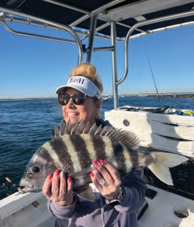 A smiling woman in a white visor and sunglasses holding up a striped sheepshead on a boat, with calm water and a distant shoreline behind her