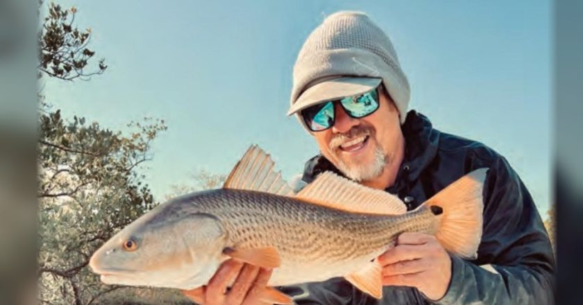 A smiling angler in a gray beanie and blue mirrored sunglasses holding a redfish in front of coastal trees under a clear blue sky