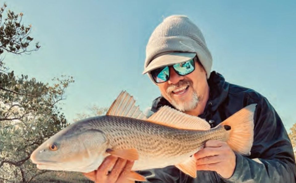 A smiling angler in a gray beanie and blue mirrored sunglasses holding a redfish in front of coastal trees under a clear blue sky