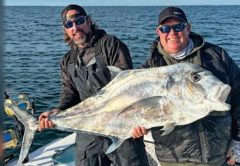 Two anglers in caps and sunglasses holding up a huge African pompano on a boat, with the open Atlantic behind them