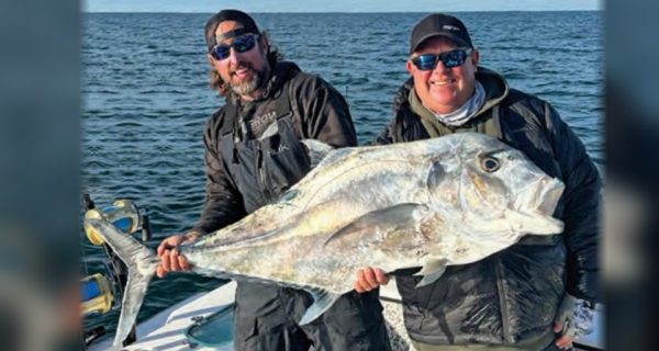 Two anglers in caps and sunglasses holding up a huge African pompano on a boat, with the open Atlantic behind them