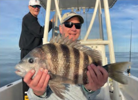 An angler in a cap and sunglasses holding up a big sheepshead on a boat, with a second angler standing behind him and calm water beyond