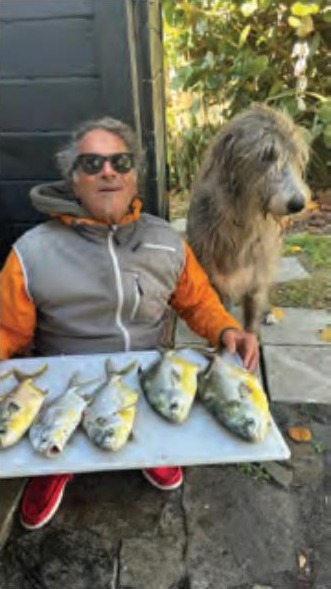Marco Pompano in sunglasses and a gray and orange jacket holding a tray of five pompano, with a big shaggy gray dog beside him