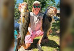 A smiling angler in a white cap and green mirrored sunglasses kneeling on the grass, holding up a snook in one hand and a largemouth bass in the other