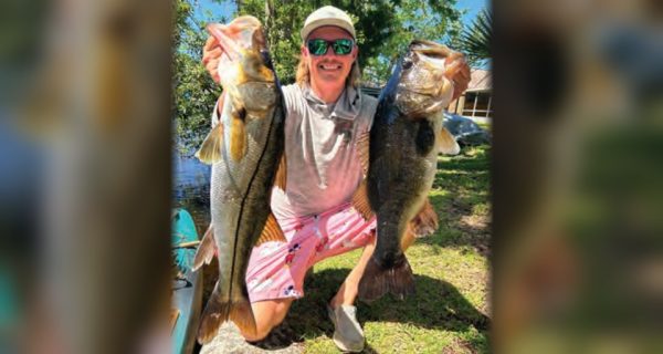 A smiling angler in a white cap and green mirrored sunglasses kneeling on the grass, holding up a snook in one hand and a largemouth bass in the other