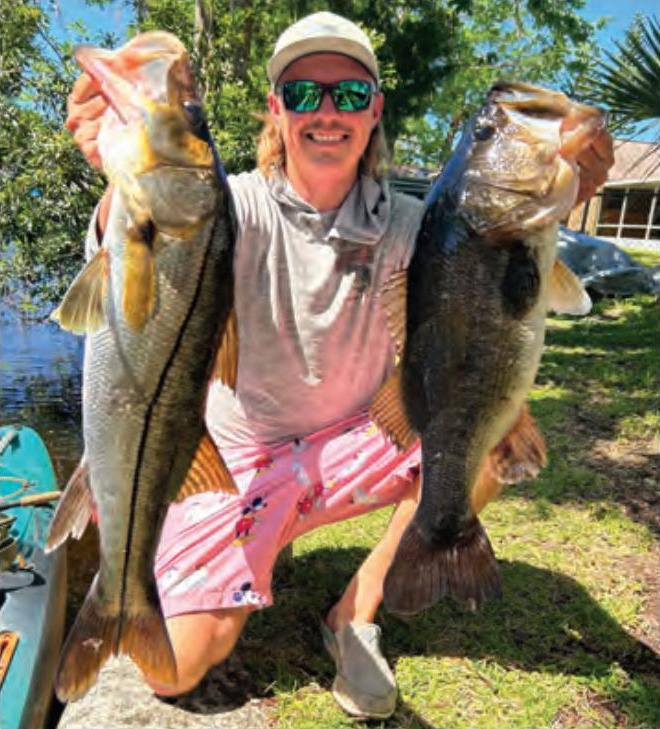 A smiling angler in a white cap and green mirrored sunglasses kneeling on the grass, holding up a snook in one hand and a largemouth bass in the other