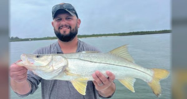 A bearded angler with sunglasses on the brim of his cap holding a snook on a boat, with open water and a tree line behind him