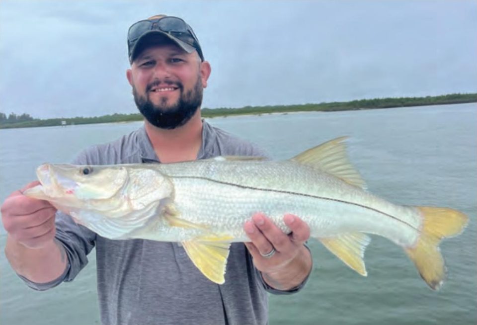 A bearded angler with sunglasses on the brim of his cap holding a snook on a boat, with open water and a tree line behind him