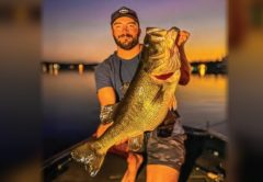Capt. Tyler Woolcott in a cap kneeling on the deck of his bass boat at sunset, holding up a giant largemouth bass