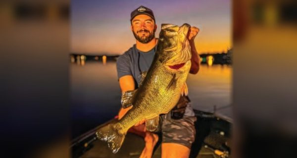 Capt. Tyler Woolcott in a cap kneeling on the deck of his bass boat at sunset, holding up a giant largemouth bass