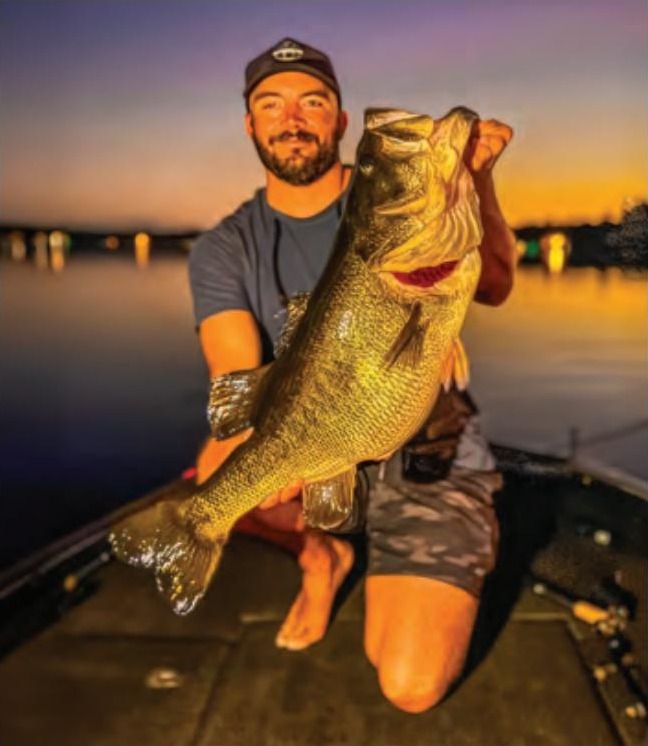 Capt. Tyler Woolcott in a cap kneeling on the deck of his bass boat at sunset, holding up a giant largemouth bass