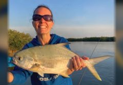 A smiling woman in sunglasses and a blue hooded sweatshirt holding up a pompano on a boat, with calm water and mangroves behind her