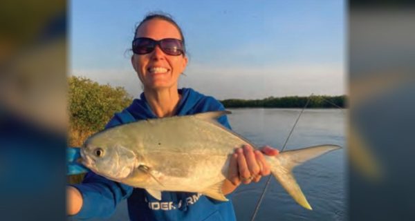 A smiling woman in sunglasses and a blue hooded sweatshirt holding up a pompano on a boat, with calm water and mangroves behind her