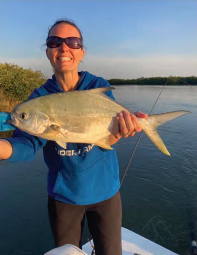 A smiling woman in sunglasses and a blue hooded sweatshirt holding up a pompano on a boat, with calm water and mangroves behind her