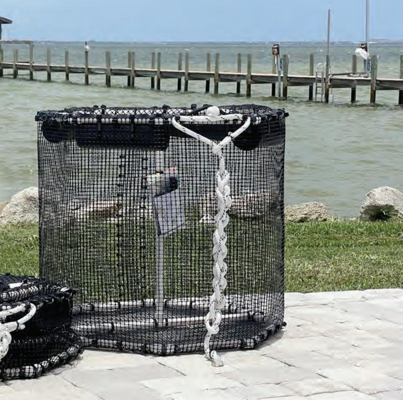 A tall black mesh bait pen with white rope handles on a waterfront patio, with a second pen beside it and a long pier behind