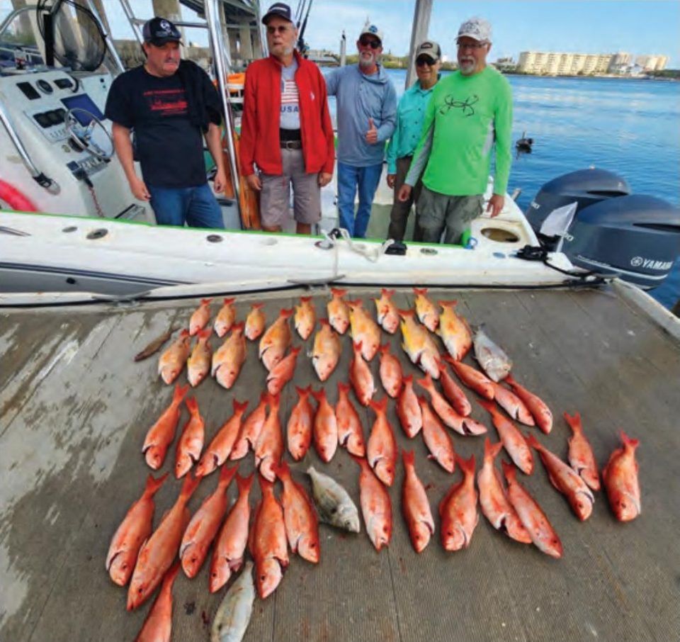 Five anglers standing on a boat at the dock behind a big spread of snapper and other bottom fish laid out on the dock