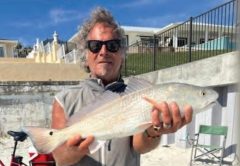 Marco Pompano in sunglasses and a gray vest holding up a redfish on the beach, with a red bike and beachfront homes behind him