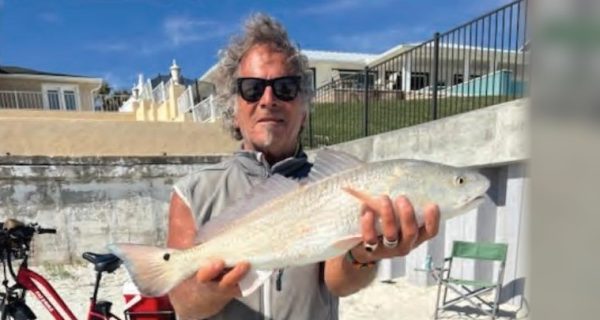 Marco Pompano in sunglasses and a gray vest holding up a redfish on the beach, with a red bike and beachfront homes behind him