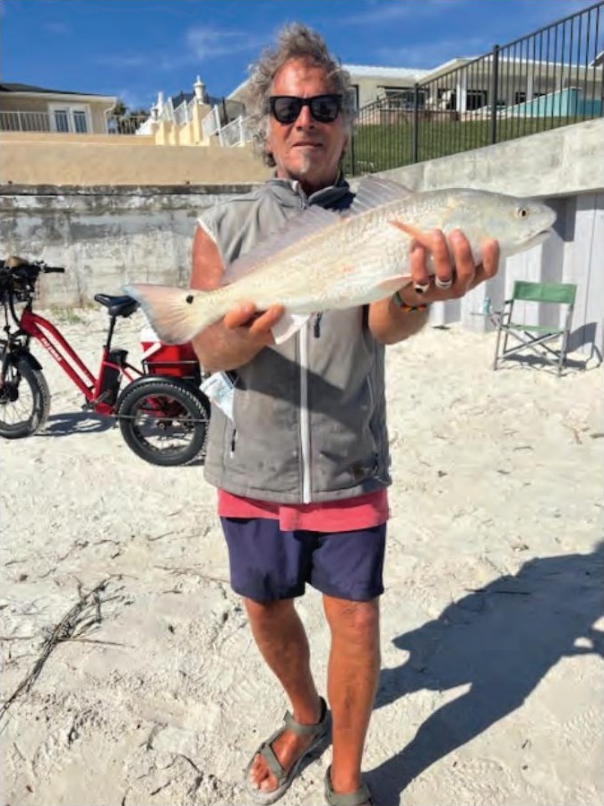 Marco Pompano in sunglasses and a gray vest holding up a redfish on the beach, with a red bike and beachfront homes behind him