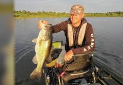 An angler in a camo cap and life vest holding up a big largemouth bass from his kayak on a lake