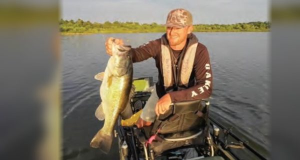 An angler in a camo cap and life vest holding up a big largemouth bass from his kayak on a lake