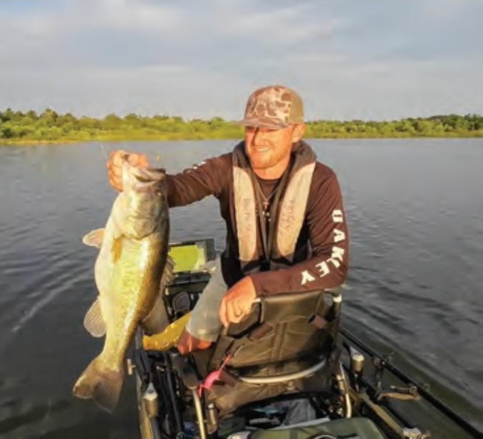 An angler in a camo cap and life vest holding up a big largemouth bass from his kayak on a lake