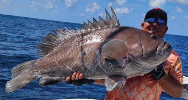 Capt. Tim Russell in a cap and sunglasses holding up a huge grouper on his boat offshore, with blue water behind him
