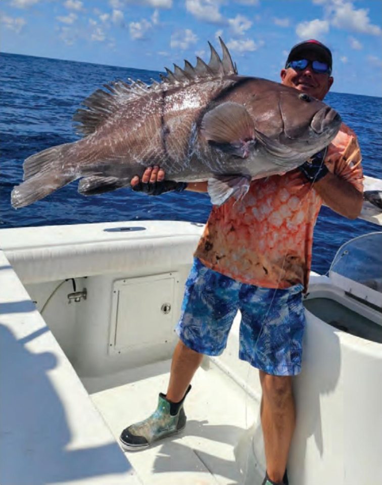 Capt. Tim Russell in a cap and sunglasses holding up a huge grouper on his boat offshore, with blue water behind him