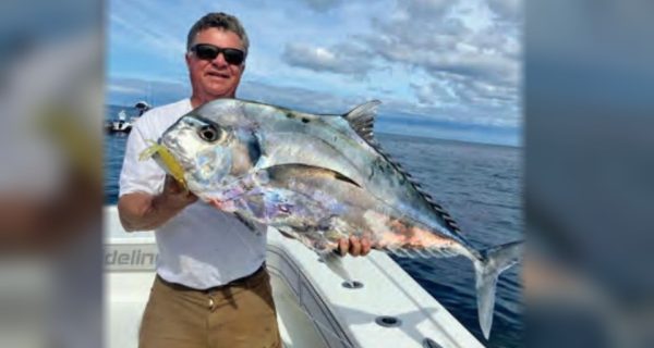An angler in sunglasses holding up a big African pompano on a boat, with blue water and clouds behind him