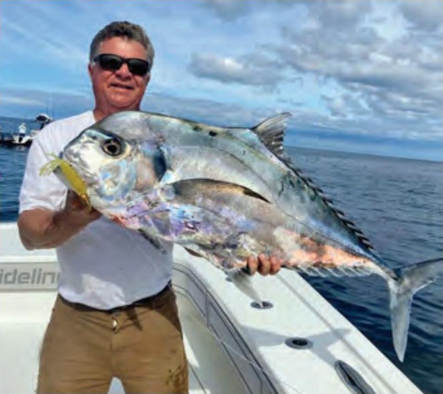 An angler in sunglasses holding up a big African pompano on a boat, with blue water and clouds behind him