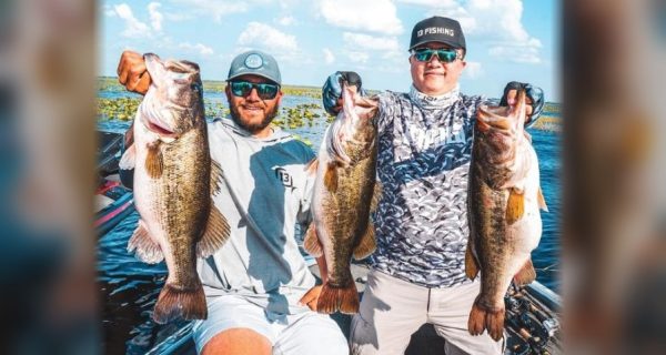Two anglers in caps and sunglasses kneeling on the deck of a bass boat, each holding up two big largemouth bass, with lily pads behind them