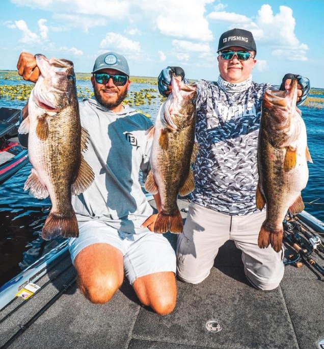 Two anglers in caps and sunglasses kneeling on the deck of a bass boat, each holding up two big largemouth bass, with lily pads behind them