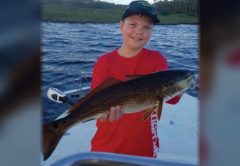A young angler in a cap and red shirt holding up a redfish on a boat, with the river and marsh behind him