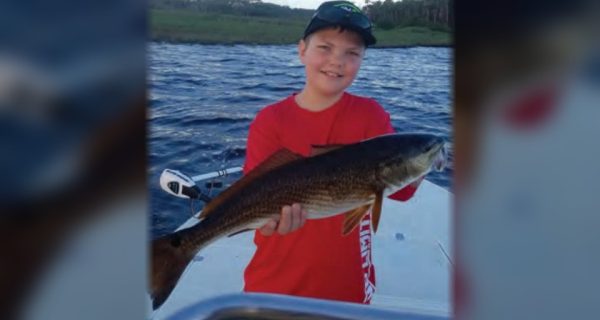 A young angler in a cap and red shirt holding up a redfish on a boat, with the river and marsh behind him