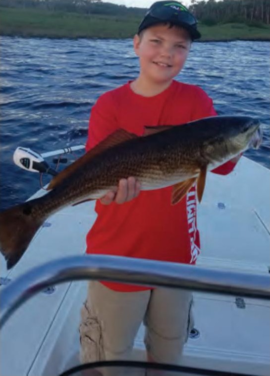 A young angler in a cap and red shirt holding up a redfish on a boat, with the river and marsh behind him