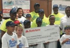 Children in Academic Anglers shirts and caps with adult volunteers, holding up a large State Farm check at a fishing clinic
