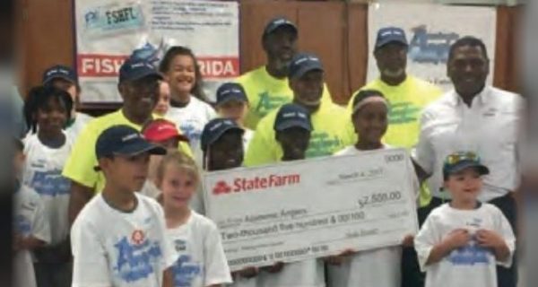Children in Academic Anglers shirts and caps with adult volunteers, holding up a large State Farm check at a fishing clinic