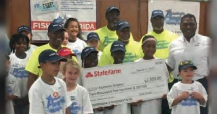 Children in Academic Anglers shirts and caps with adult volunteers, holding up a large State Farm check at a fishing clinic