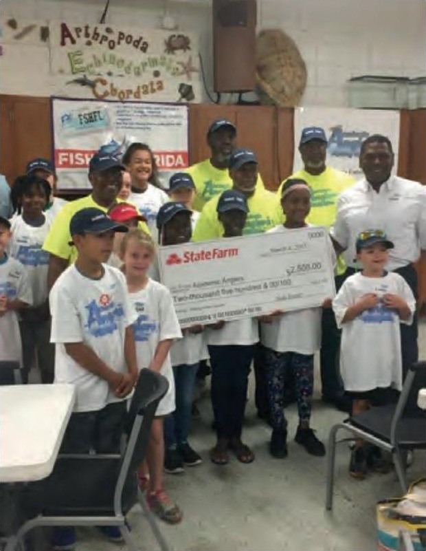 Children in Academic Anglers shirts and caps with adult volunteers, holding up a large State Farm check at a fishing clinic