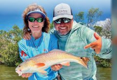 A smiling woman in a visor and a man in a Starke cap, both in sunglasses, holding a redfish in front of the mangroves