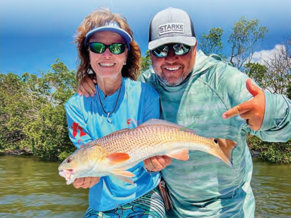 A smiling woman in a visor and a man in a Starke cap, both in sunglasses, holding a redfish in front of the mangroves
