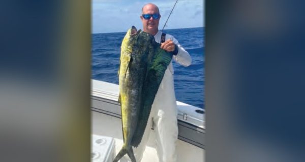 Capt. Scott Housel in sunglasses holding up a big mahi on his boat offshore, with blue water behind him