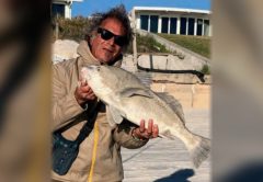 Marco Pompano in sunglasses holding up a black drum on the beach, with beachfront homes behind him