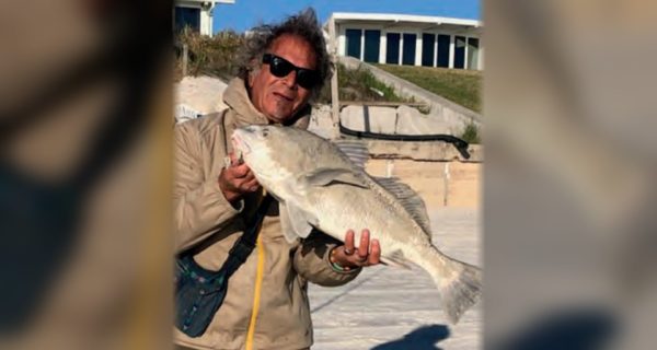 Marco Pompano in sunglasses holding up a black drum on the beach, with beachfront homes behind him