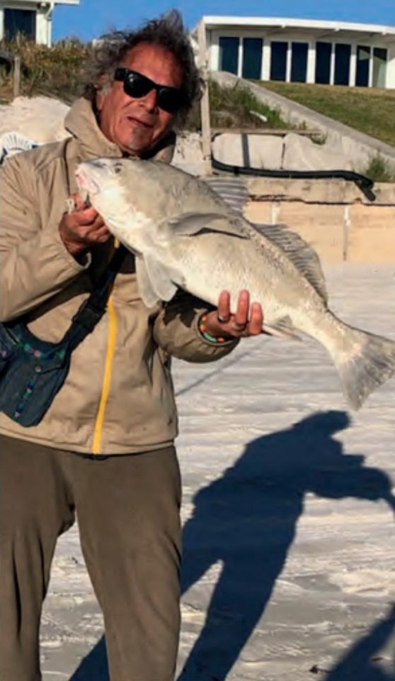 Marco Pompano in sunglasses holding up a black drum on the beach, with beachfront homes behind him