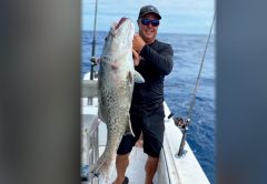 Capt. Scott Housel in a cap and sunglasses holding up a big grouper on the deck of his boat offshore