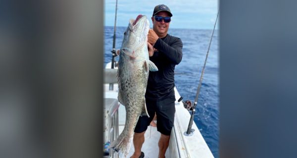 Capt. Scott Housel in a cap and sunglasses holding up a big grouper on the deck of his boat offshore