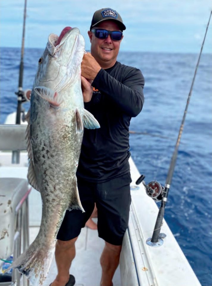 Capt. Scott Housel in a cap and sunglasses holding up a big grouper on the deck of his boat offshore